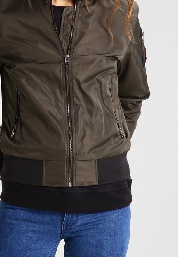 Personne portant une veste bomber vert olive avec poches zippées et un t-shirt noir en dessous, associée à un jean bleu.