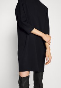 Robe noire surdimensionnée avec des manches trois-quarts, faite d'un tissu doux. Le design présente un col rond et des fentes latérales discrètes.