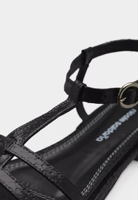 Μαύρο επίπεδο σανδάλι με υφή, λουράκι αστραγάλου και ασημένια αγκράφα, με ραμμένη πάτο και λουριά που διασταυρώνονται πάνω από το πόδι.