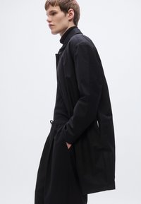 Μαύρο μακρύ παλτό από λείο ύφασμα, με μινιμαλιστικό σχέδιο, άνετη εφαρμογή και πλαϊνές τσέπες. Φοριέται πάνω από στενό μαύρο τοπ και παντελόνι.