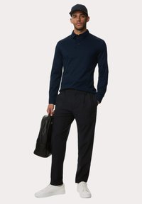 Marineblaues Langarm-Polohemd, schwarze Hose, weiße Sneakers, hält einen schwarzen Rucksack. Verfügt über einen klassischen Kragen und ein enganliegendes Design.