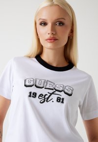 Biela tričko s čiernym rebrovaným výstrihom a potlačou loga "GUESS est. 1981" v čiernej farbe, s tučným písmom a elegantným dizajnom.