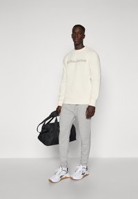 Cremefarbene Sweatshirt mit "BJÖRN BORG"-Aufdruck, graue Sweatpants und weiße Sportschuhe mit schwarzen Akzenten. Trägt eine schwarze Sporttasche.