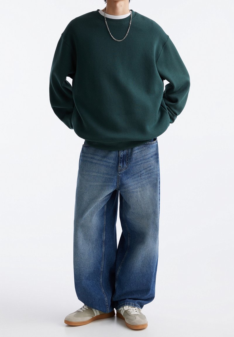 Personne portant un sweat-shirt oversize vert foncé, un jean bleu à jambes larges, des baskets beige et un collier chaîne argenté, debout les mains derrière le dos.