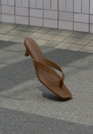 Sandalia de tacón marrón de cuero Steve Madden flotando sobre suelo pavimentado con pared de azulejos al fondo.