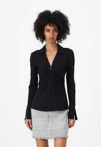 Maglietta nera a maniche lunghe con un design arricciato verticale, colletto a punta e dettaglio con zip sullo scollo. Tessuto liscio e vestibilità sartoriale.