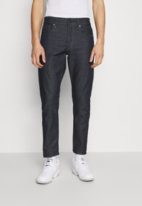 Dark denim jeans with a slim fit, front pockets, and a button closure. Worn with white sneakers, highlighting the fabric's smooth texture.