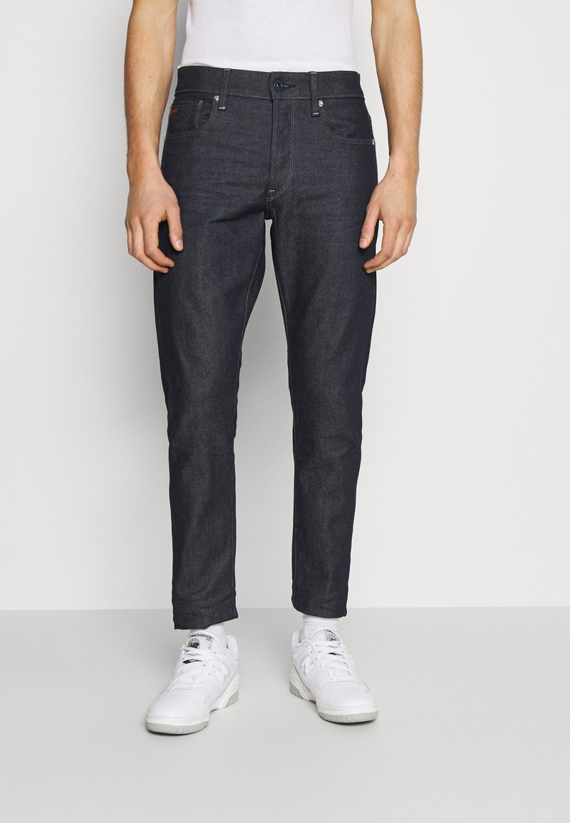 Dark denim jeans with a slim fit, front pockets, and a button closure. Worn with white sneakers, highlighting the fabric's smooth texture.