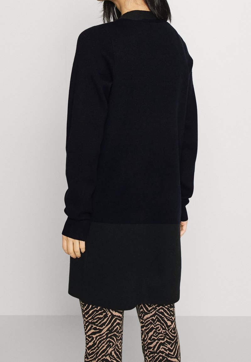 Femme portant un long pull noir et des leggings à motifs, debout dos à la caméra sur un fond uni.
