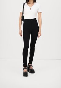 Jeans negros de cintura alta con un ajuste ceñido, combinados con una blusa blanca de manga corta con escote en forma de ojo de cerradura y zapatos de plataforma negros y gruesos.