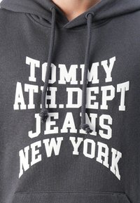 Pilka dėvima su gobtuvu su baltu užrašu „TOMMY ATH.DEPT JEANS NEW YORK“. Yra su raišteliu ir minkšta, tekstūruota medžiaga.
