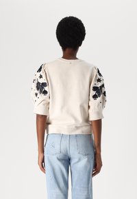 Crème-kleurige sweatshirt met geborduurde zwarte bloemenpatronen op de poffende mouwen, gecombineerd met lichtblauwe jeans met steraccenten.