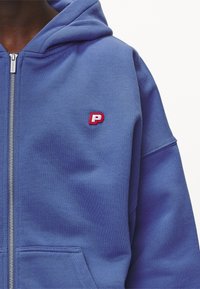Felpa con cappuccio in tessuto azzurro chiaro, dotata di una cerniera frontale e di un piccolo logo rosso ricamato "P" sul petto.