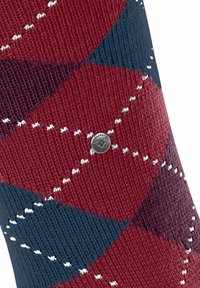 Nahaufnahme eines gemusterten Gewebes mit roten, blauen und dunkelvioletten Rauten, weißen Nähten und einem kleinen runden Logo.