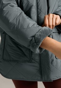 Osoba podvija rukav tamnosive jakne s prošaranim uzorkom, unutar kojeg se nalazi mekani sivi umjetni krzno, pokazujući dio podlaktice.