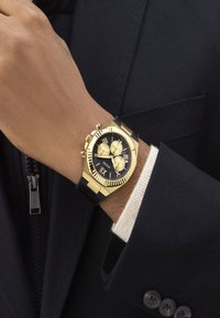 Montre chronographe Guess noire et dorée avec des chiffres romains portée au poignet, sur une manche blanche et une veste de costume sombre.