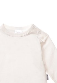 Langärmliges cremefarbenes Baumwollhemd mit rundem Halsausschnitt und drei Druckknöpfen an einer Schulter. Weiche Textur mit glatter Oberfläche.