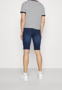 Hombre vistiendo una camiseta de rayas blancas y negras, pantalones cortos de mezclilla azul oscuro, calcetines blancos y zapatillas blancas, de pie contra un fondo sencillo.