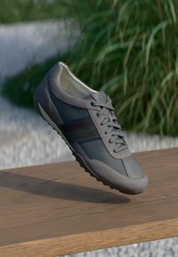 Zapatilla deportiva gris que presenta una combinación de materiales lisos y texturizados, con un diseño de cordones y suela de goma, situada sobre un fondo natural.
