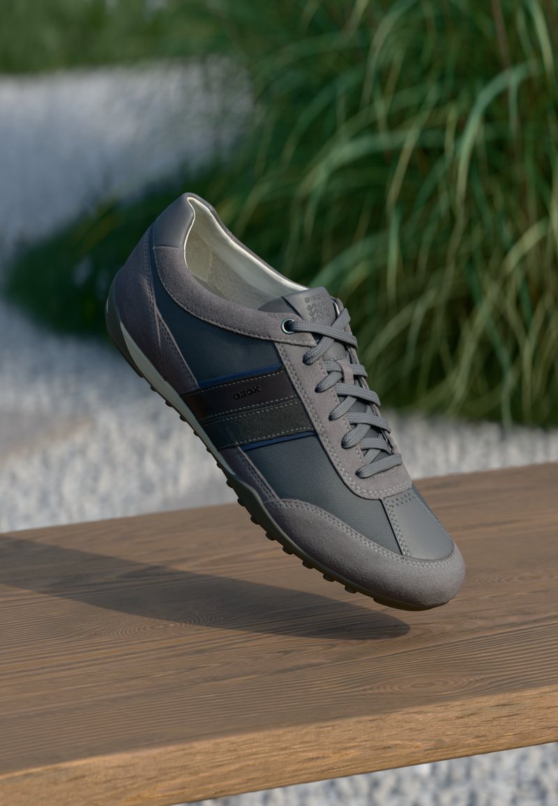 Zapatilla deportiva gris que presenta una combinación de materiales lisos y texturizados, con un diseño de cordones y suela de goma, situada sobre un fondo natural.