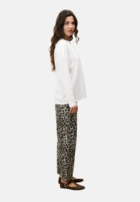 Vrouw met lang golvend haar, gekleed in een losse witte blouse met lange mouwen, een broek met luipaardprint en bruine platte schoenen met een gesp, staande in profiel.