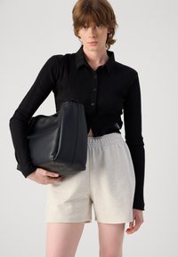 Svart ribbestrikket skjorte med knapper og lange ermer, parret med lysegrå bomullsshorts. Holder en svart lærveske med en strukturert design.
