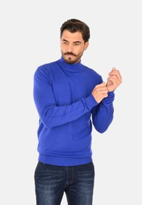 Maglione dolcevita blu in tessuto a maglia liscia, design aderente, polsini e orlo a coste, abbinato a jeans scuri in denim.