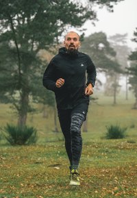Sort let løbejakke, leggings med camouflage-mønster og grønne sko. Løb på græs med træer i baggrunden under tågede forhold.