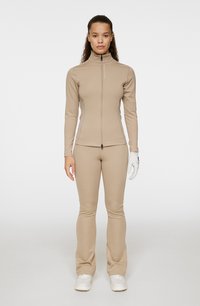 Beige sportig zip-up topp och utsvängda byxor av slät tyg, i kombination med en vit handske och skor, med minimal utsmyckning.