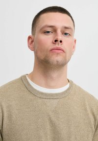 Beige gebreide trui met een ronde halslijn, textuurstof en subtiele details op de schouders. Draag het over een witte T-shirt.