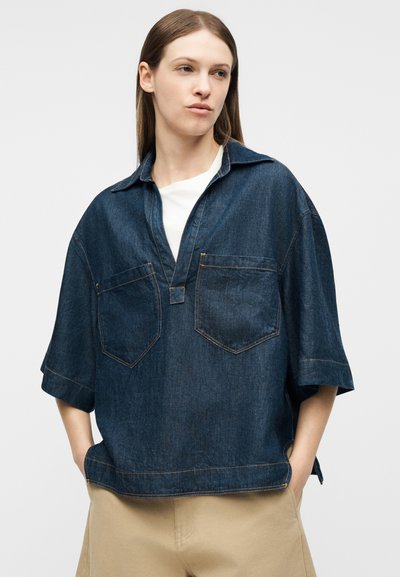 Νέα γυναίκα που φοράει μπλε σκούρο τζιν πουκάμισο oversized πάνω από λευκό τοπ, μπεζ παντελόνι, με τα χέρια στις τσέπες, κοιτάζοντας πλάγια.