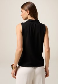 Top senza maniche nero con colletto, realizzato in un tessuto liscio. Il retro presenta una leggera drappeggiatura, abbinato a pantaloni di colore chiaro.