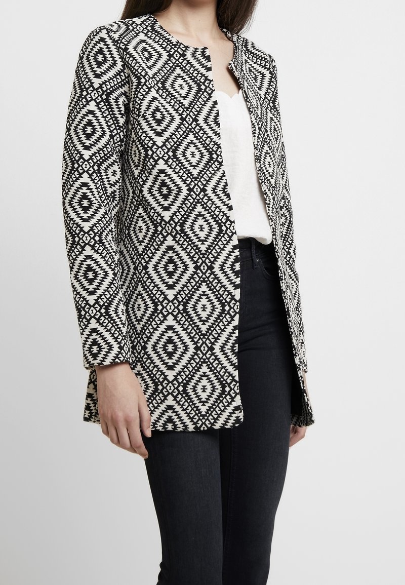 Femme portant une veste à manches longues à motif géométrique noir et blanc sur un haut blanc et un pantalon noir, sur un fond uni.
