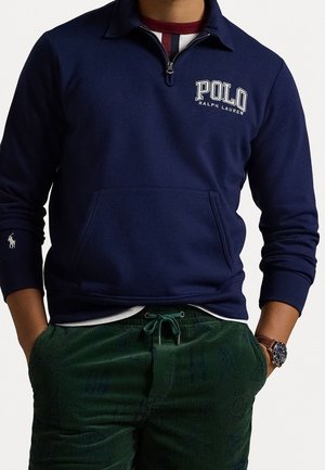Mężczyzna w granatowym swetrze Polo Ralph Lauren z zamkiem na ćwierć długości i ciemnozielonych spodniach sztruksowych, ręce w kieszeniach.