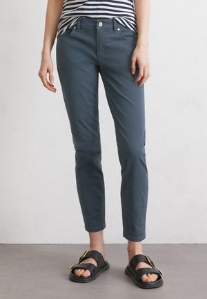 Persoon in teal slim-fit broek, gestreepte top ingestopt, en zwarte open-toe sandalen met gouden gespen, staand op een grijze vloer.
