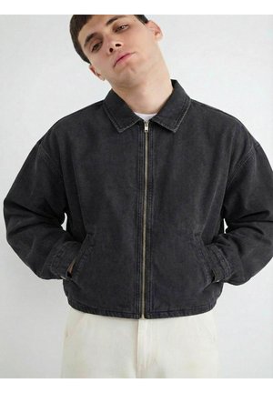 Mladý muž v čiernej bunde so zipsom a golierom, rukami v vreckách a bielych nohaviciach na jednoduchom svetlom pozadí.