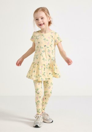 Smilende ung pige i gul blomsterkjole og matchende leggings, iført hvide sneakers, stående mod en enkel lys baggrund.