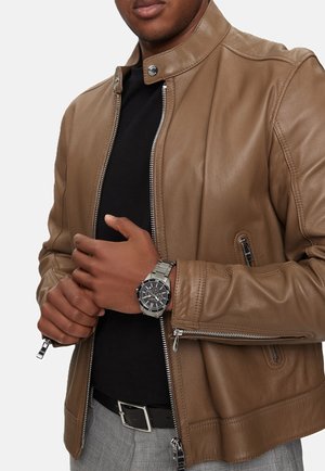 Rudas odinė striukė su aukštu apykakle, užtrauktuku priekyje ir šoninėmis kišenėmis; dėvima ant juodos marškinėlių. Ranke yra sidabrinis laikrodis.