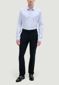 Uomo che indossa una camicia da uomo azzurra chiara con bottoni, pantaloni neri eleganti e mocassini neri lucidi, in piedi davanti a uno sfondo neutro.