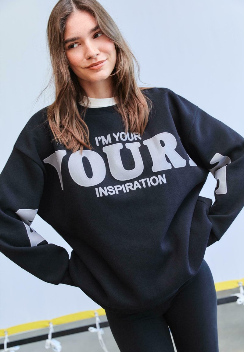 Čierna mikina s bielym tučným textom "JA SOM TVOJ" a "INŠPIRÁCIA", s uvoľneným dizajnom a dlhými rukávmi.