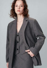 Blazer grigio a righe con revers a punta e taschino sul petto, abbinato a un gilet coordinato con bottoni neri. Tessuto dalla texture liscia.