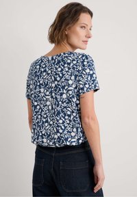 Top corto blu navy con motivo floreale bianco, vestibilità morbida, scollatura rotonda e tessuto morbido; abbinato a jeans scuri.