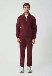 Uomo in piedi che indossa un maglione maroon con mezza zip e pantaloni jogger abbinati, con scarpe da ginnastica bianche su uno sfondo semplice.