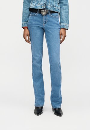 Person iført lyseblå jeans med rette ben, svarte spisse støvler, en denimjakke og et svart belte med en stor sølvspenne formet som et hjerte.