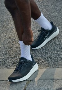 Schwarze Sportschuhe mit strukturiertem Obermaterial, weißer Zwischensohle und schwarzen Schnürsenkeln. Getragen mit weißen gerippten Socken. Hintergrund aus Asphalt im Freien.