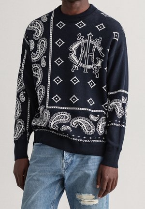 Homme portant un pull bleu marine avec des motifs blancs paisley et en losange et un jean bleu clair déchiré, debout devant un fond uni.