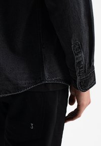 Zwarte denimjas met een knoopsluiting aan de manchetten, voorzien van ton-sur-ton stiksels en een label met een "J" aan de zijkant. De stof heeft een gestructureerd uiterlijk.