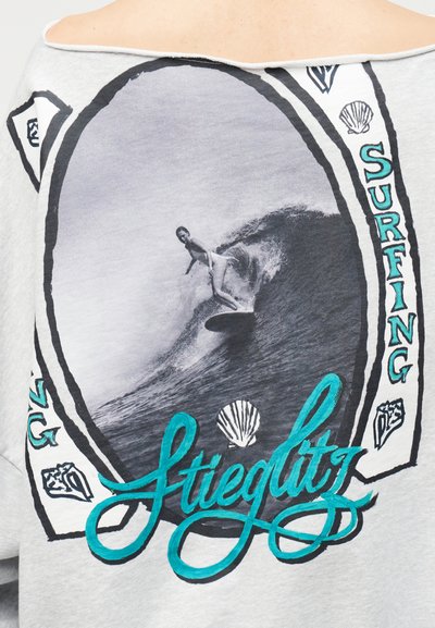 Σερφίστερ που καβαλάει ένα μεγάλο κύμα μέσα σε οβάλ πλαίσιο, με το κείμενο "Surfing" και εικονογραφήσεις κοχυλιών σε γκρι υφασμάτινο φόντο.