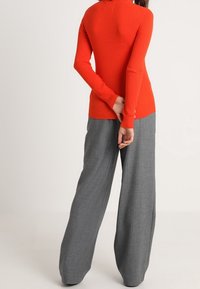 Top rouge à manches longues côtelées avec un col montant, associé à un pantalon gris à jambes larges en tissu texturé. Vue de dos affichée.