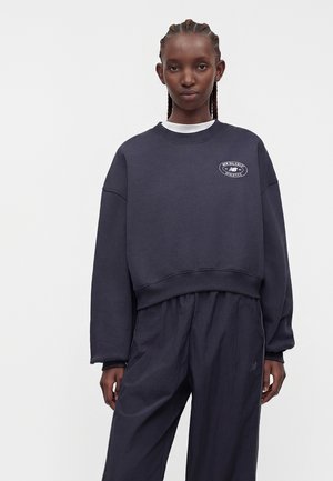 Vrouw met gevlochten haar die een donkerblauwe sweatshirt met het New Balance Athletics-logo en bijpassende losse broek draagt, staand tegen een effen achtergrond.